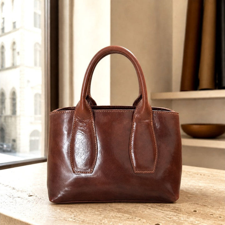A brown leather handbag with double top handles, standing on a stone ledge in front of a window | Artisans