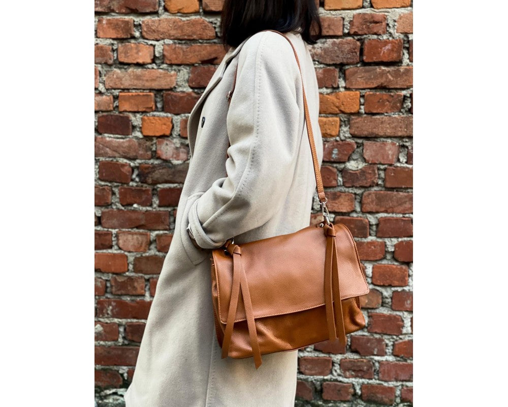 A woman in a light beige coat carries a tan leather shoulder bag with a flap front and long strap against a red brick wall background.