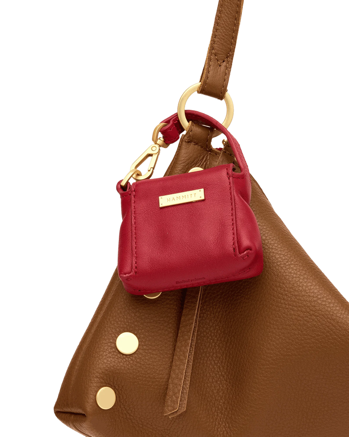 A close-up of a brown pebbled leather handbag with a small red leather shoulder bag charm featuring a gold-tone brand plaque.
