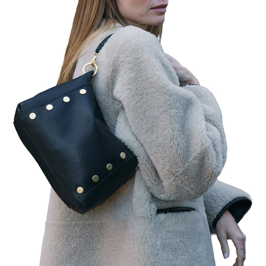 A woman wearing a light beige sherpa coat carries a black pebbled leather bucket bag with gold-tone studs and a gold ring detail on her shoulder, set against a plain white background.