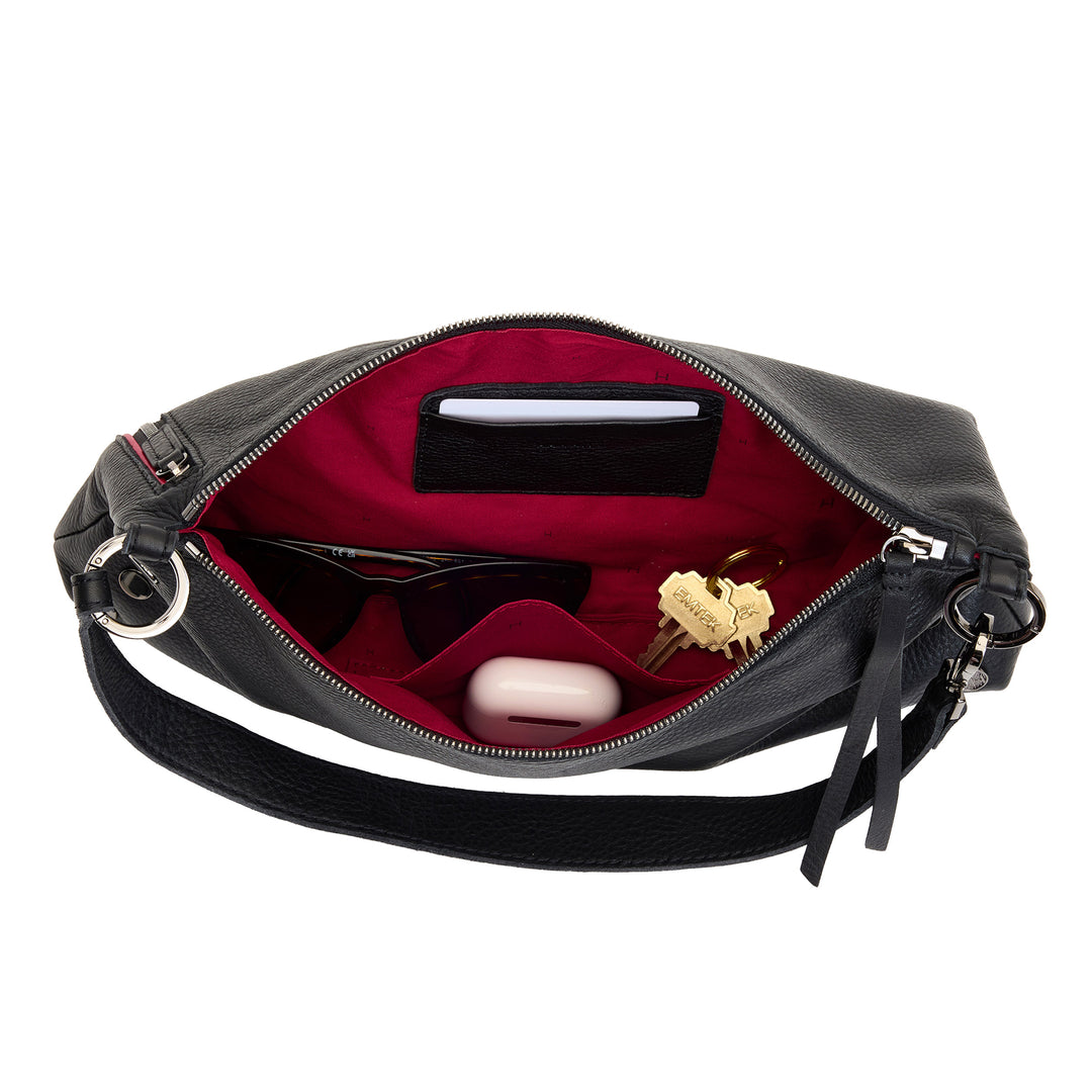 An open black pebbled leather shoulder bag with a bright red interior lining, shown against a white background. The bag contains various accessories including sunglasses, a pink case, and keys.