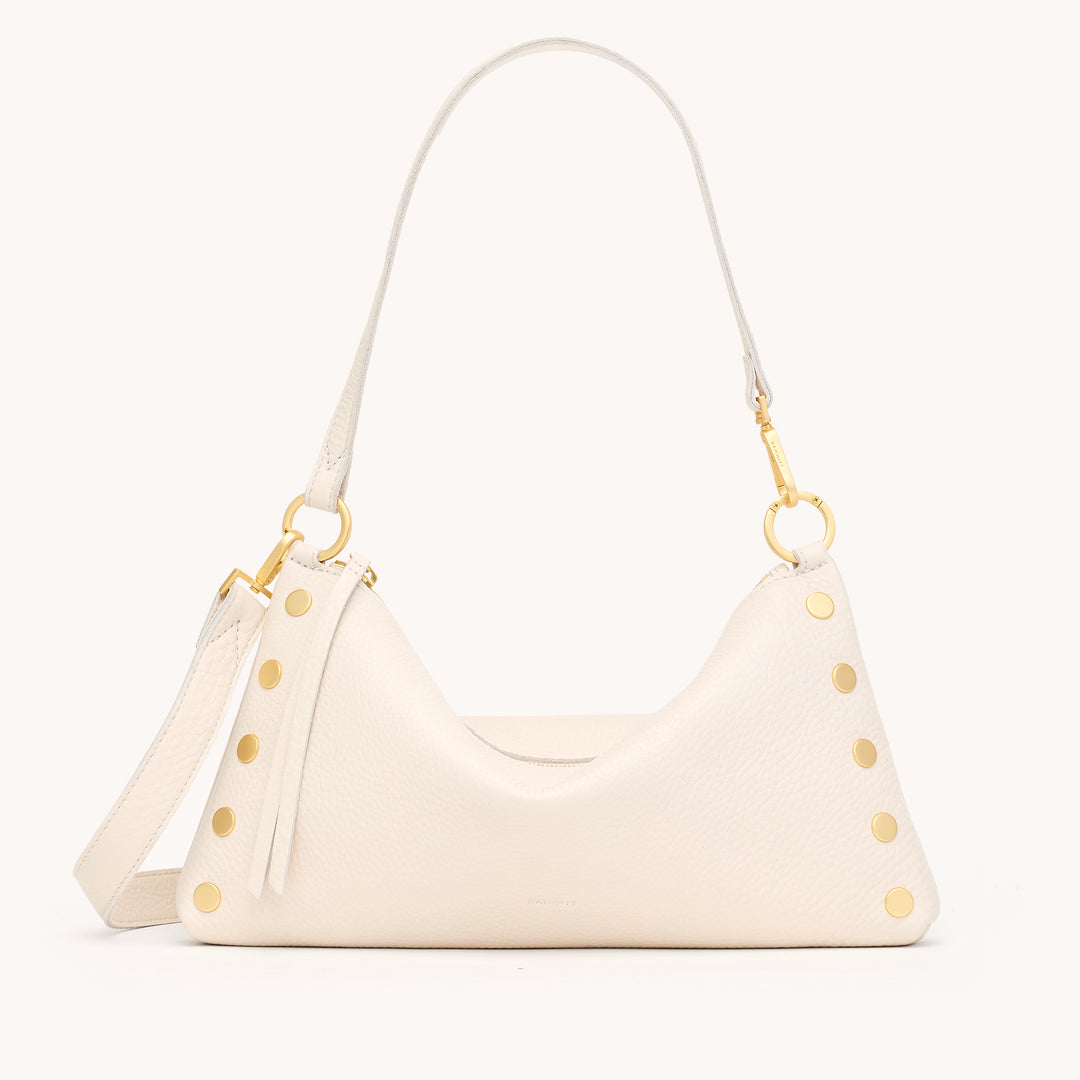 An off-white pebbled leather shoulder bag with gold-tone hardware, featuring a crescent-shaped body, a tassel detail, and a matching shoulder strap, shown against a solid off-white background.