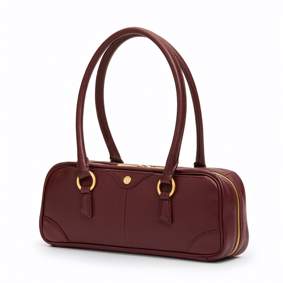 A burgundy leather handbag with gold-tone hardware, featuring dual top handles, a zippered closure, and a small round gold button on the front, set against a plain white background.