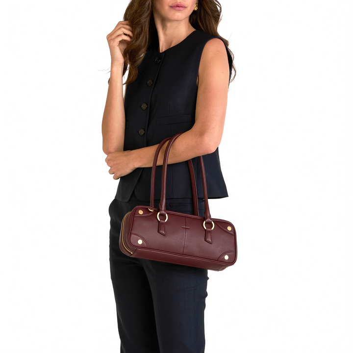 A woman wearing a sleeveless black vest and dark pants carries a burgundy leather handbag with gold-tone hardware against a plain white background.