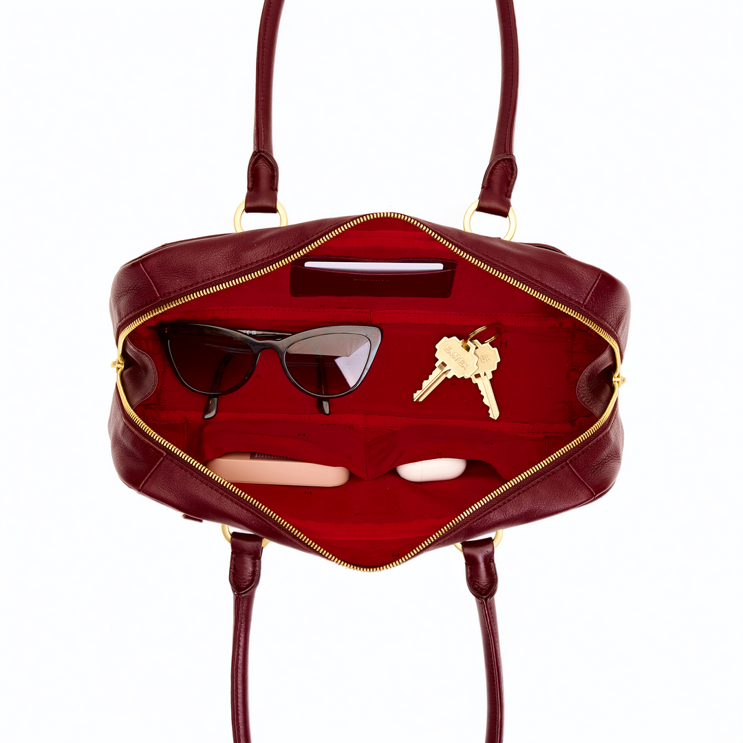 An open burgundy leather handbag with gold-tone hardware, shown from above to display its interior compartments. Inside, a pair of black sunglasses, a set of keys, and two small rectangular items are arranged on a red lining.