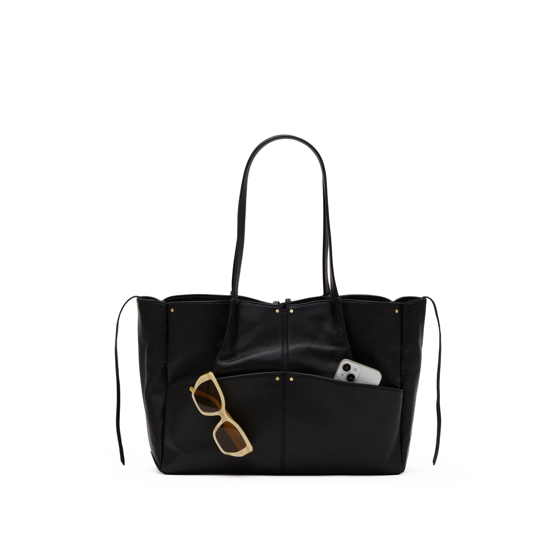 A black leather tote bag with long shoulder straps, a front slip pocket holding gold-framed sunglasses and a white phone, shown against a plain white background.