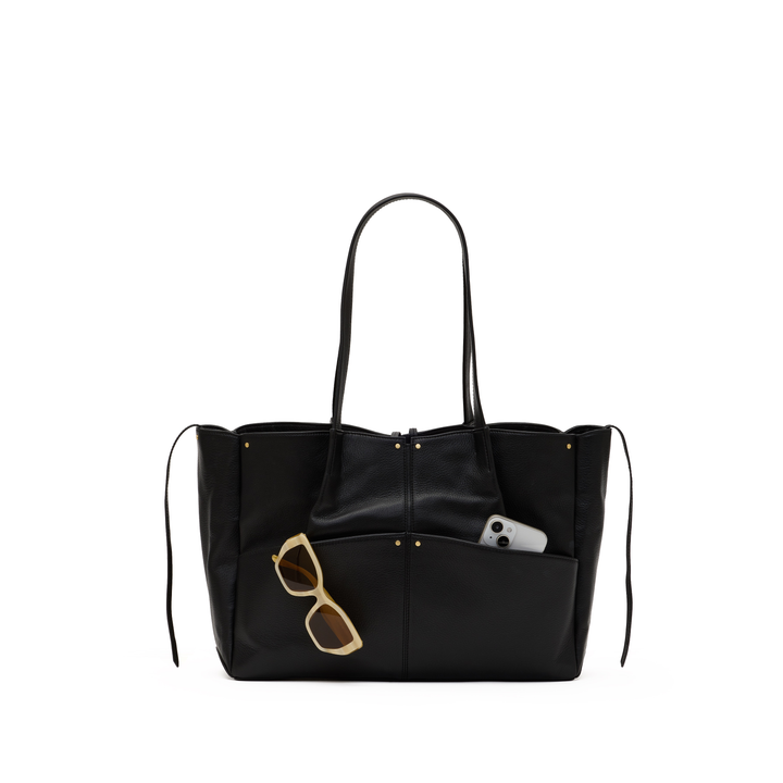A black leather tote bag with long shoulder straps, a front slip pocket holding gold-framed sunglasses and a white phone, shown against a plain white background.