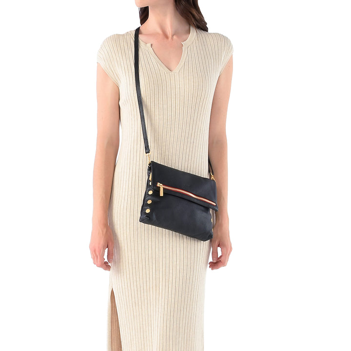 A woman wearing a cream ribbed knit dress models a black leather crossbody clutch bag with gold hardware and a thin orange-brown leather accent stripe.