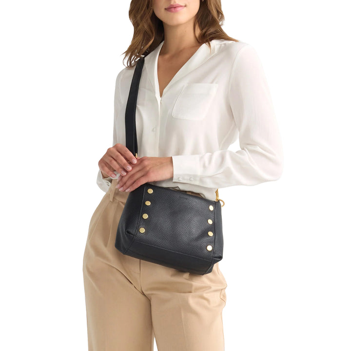 A woman wearing a white blouse and beige pants carries a black pebbled leather crossbody bag with gold-tone studs and a black strap against a plain white background.