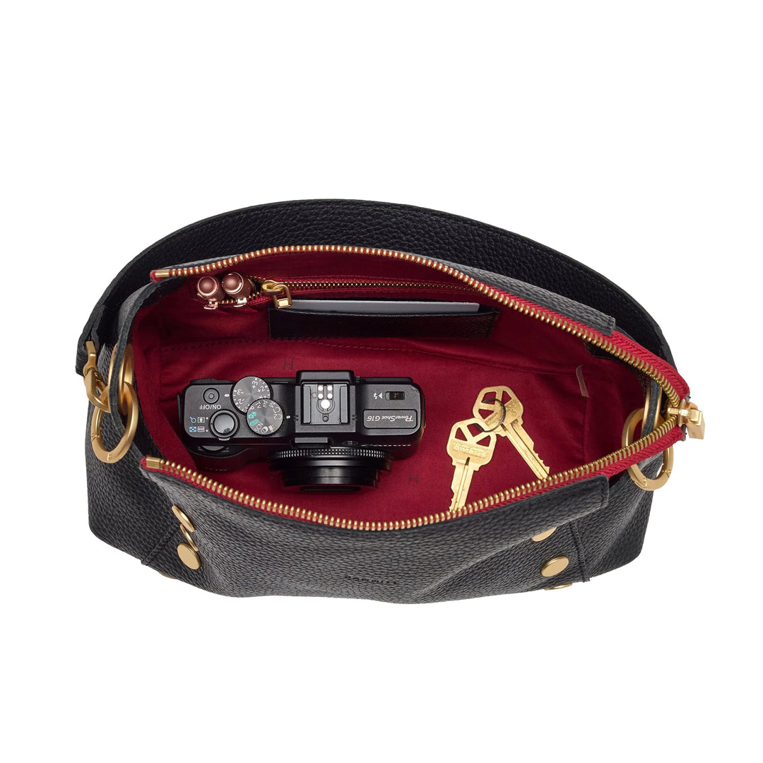 An open black pebbled leather handbag with gold-tone hardware, showing a deep burgundy red interior. Inside, a black camera, a slim black case, and a pair of gold keys are arranged.