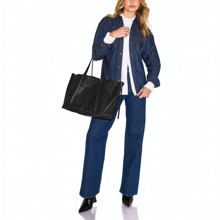 A model wearing a dark blue denim jacket, matching wide-leg jeans, a white collared shirt, and black pointed shoes, carrying a large black leather tote bag with gold-tone studs against a white studio background.
