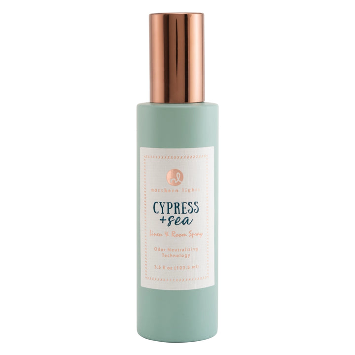 A bottle of Northern Lights Cypress + Sea linen and room spray, featuring a light sage green bottle and a metallic rose gold cap, set against a plain white background.