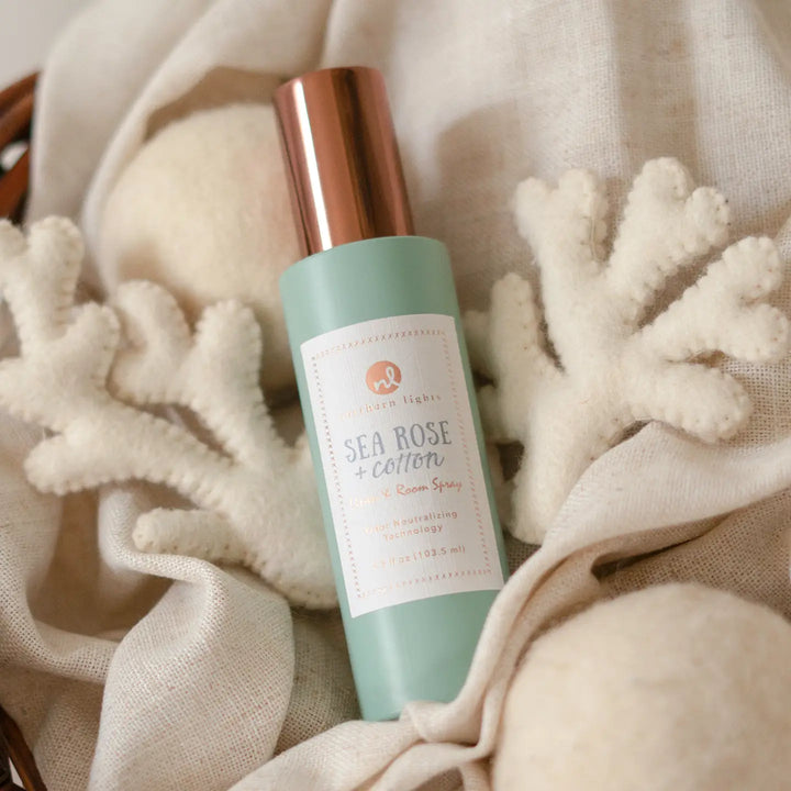 A frosted mint green bottle of Ocean Lights Sea Rose + Cotton Room Spray with a metallic rose gold cap, nestled in soft beige linen and white coral decorations.