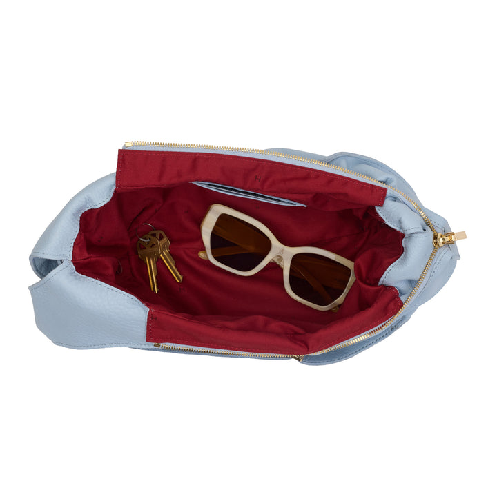 An open light blue hobo bag with a deep red interior, shown on a white background. Inside, white-framed sunglasses and a set of keys are placed as styling props.