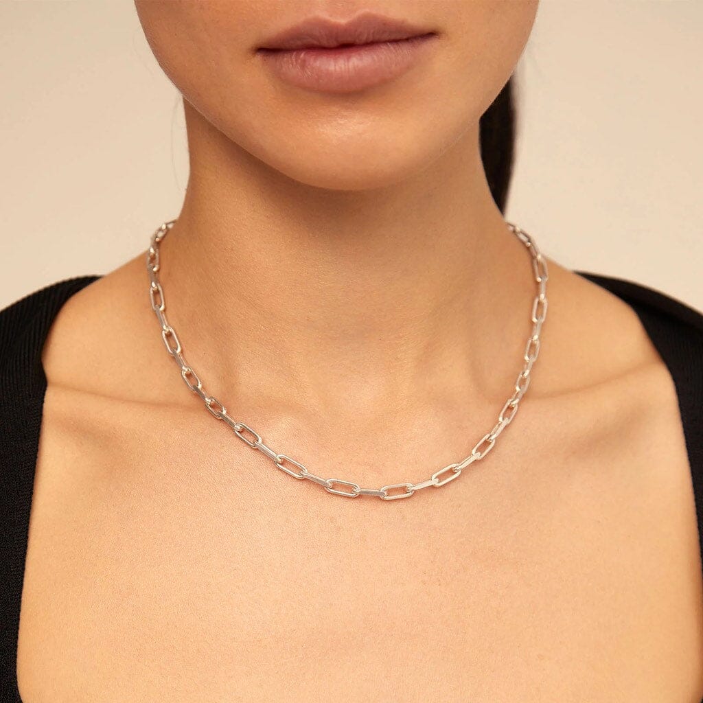 A close-up of a model wearing a silver paperclip chain necklace against a warm beige background.