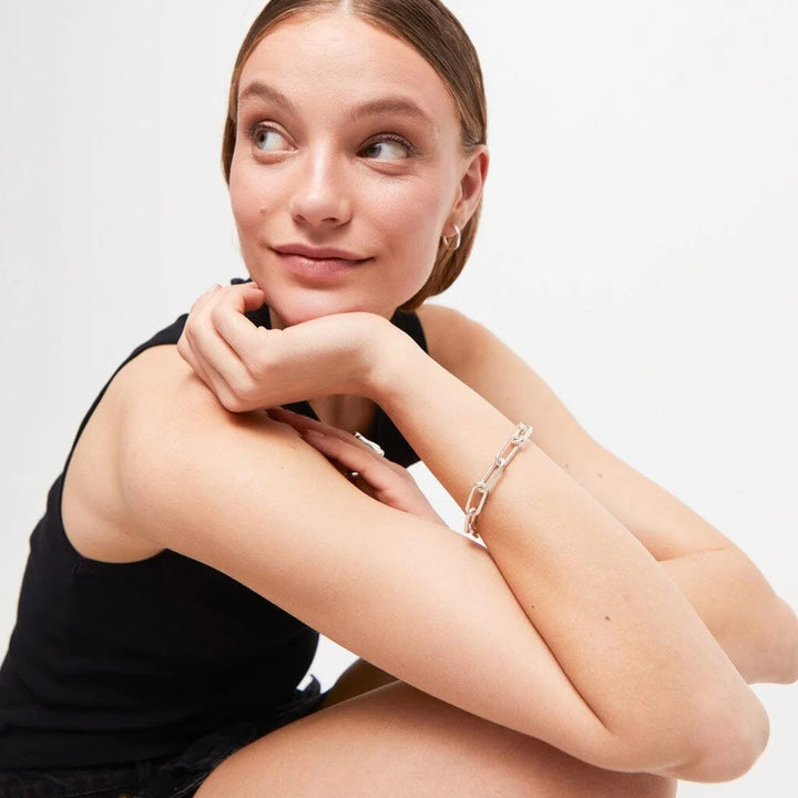A woman wearing a delicate silver chain link bracelet, posing with her arms crossed against a clean white background.
