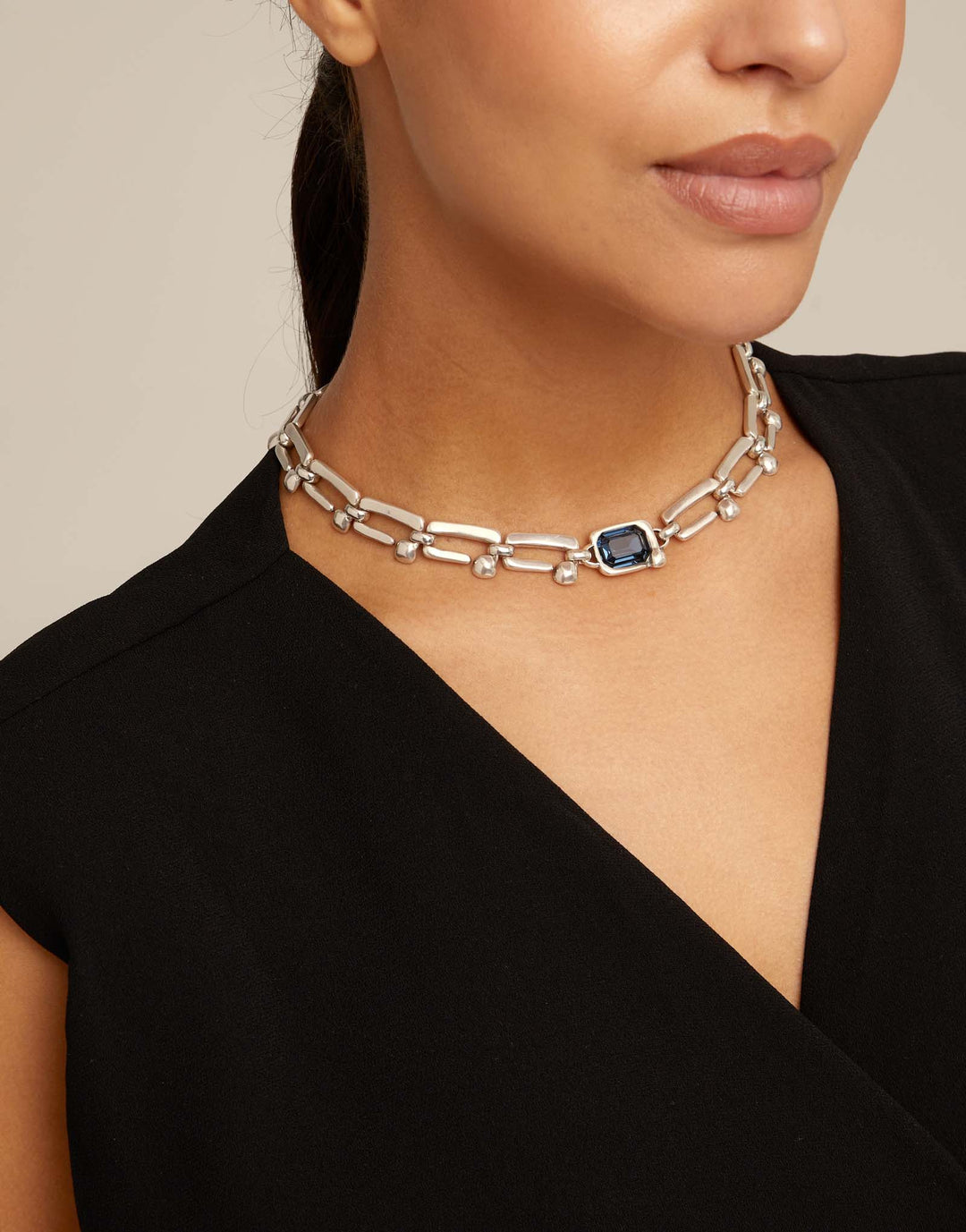 A close-up of a model wearing a silver-toned chain choker necklace with a central rectangular dark blue stone and small clear accent stones, paired with a black top.